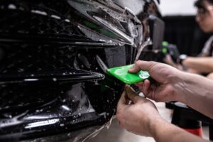 Car detailing studio employee, a wrapper, is protecting the bumper of a black car with a colorless protective PPF film.