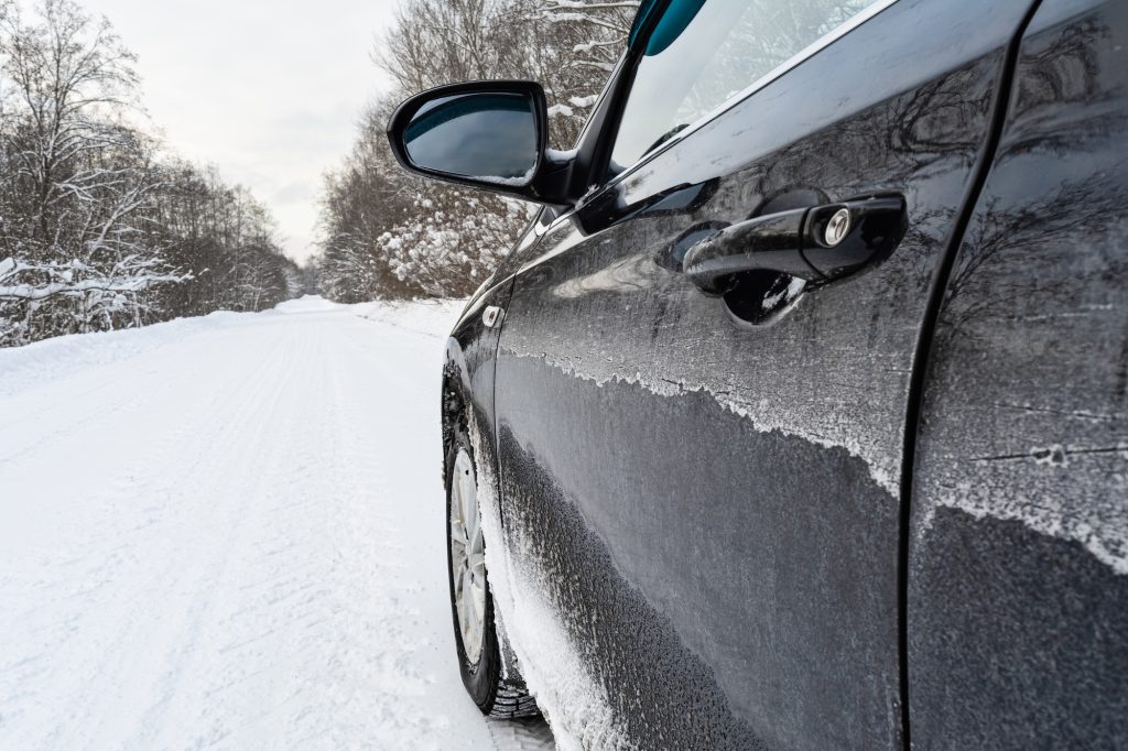 A black car on a snowy rural road. The side of the car is covered in snow and road de-icing chemicals.