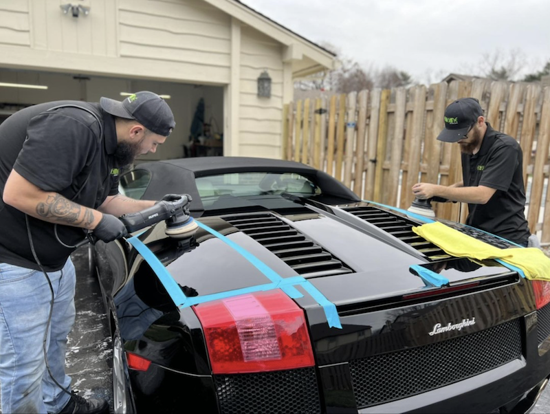 Two men detail a black Lamborghini, polishing the rear with a power buffer while blue tape marks panels for protection.